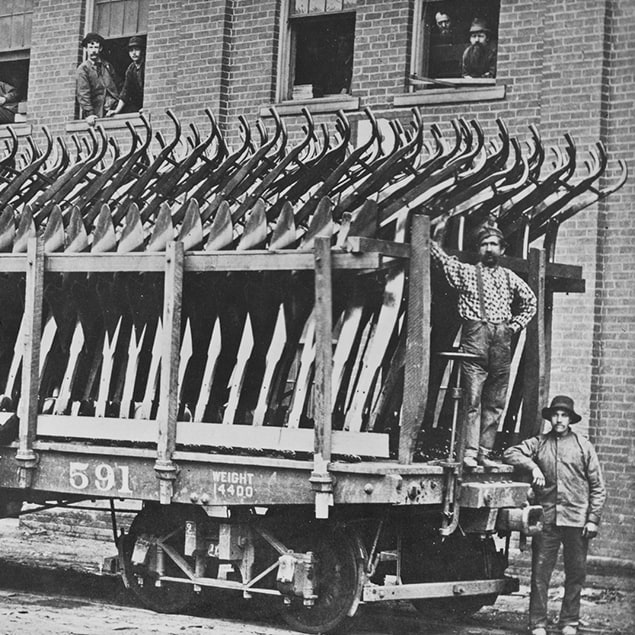 1882 рік — фотографія трьох чоловіків, які стоять біля вагону Deere & Co, завантаженого готовими до відправлення сталевими плугами, і працівників заводу, які визирають із вікон розташованої позаду будівлі.