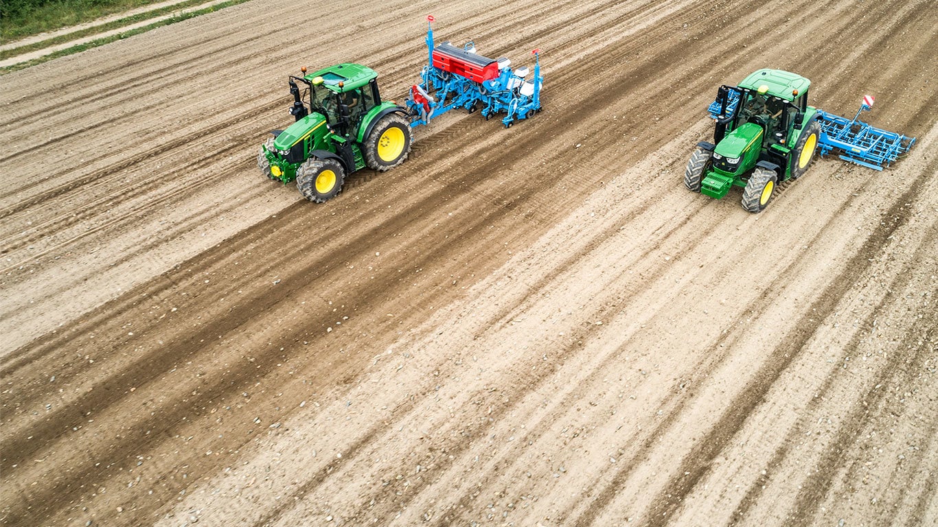 Два трактори 6M від John Deere на полі з необробленою землею. Один із них оснащено сівалкою Monsomen. Інший тягне посівний комплект Lemken.