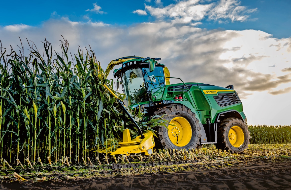 Кормозбиральний комбайн John Deere збирає кукурудзу в полі під блакитним небом із хмарами.
