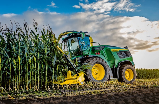 Кормозбиральний комбайн John Deere збирає кукурудзу в полі під блакитним небом із хмарами.