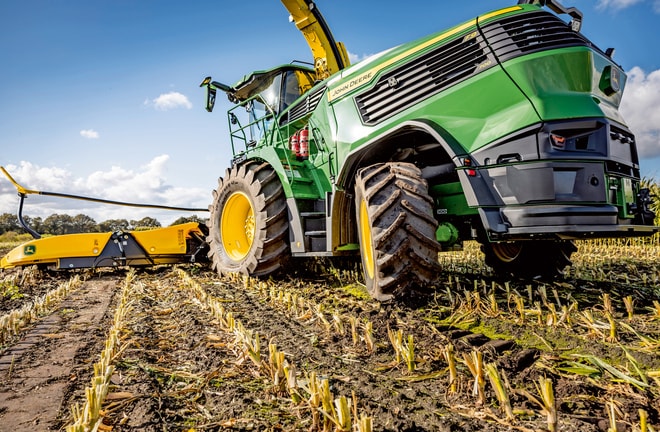 Кормозбиральний комбайн John Deere працює на кукурудзяному полі, де вже зібрано врожай, демонструючи великі шини та ріжучий механізм машини на тлі чистого блакитного неба.